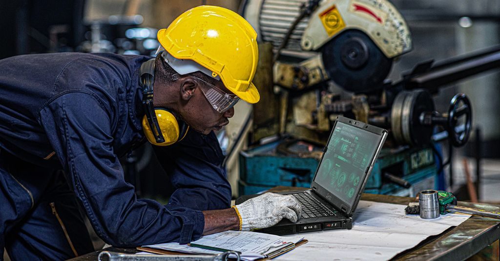 Construction worker looking at building blueprints and checking digital plans on a rugged laptop.