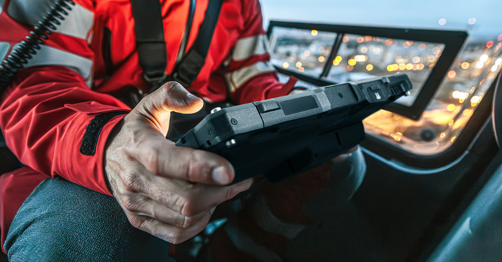 Emergency responder holding a rugged tablet inside an aircraft cockpit at dusk, with city lights visible through the window.