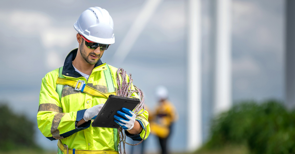 Wind farm technician wearing safety gloves uses a rugged Getac tablet to log field data near wind turbines.