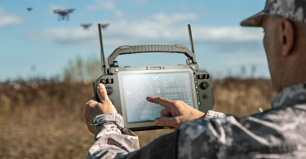 Operator in camouflage using a rugged tablet ground control station with handles and antennas to monitor drones in flight