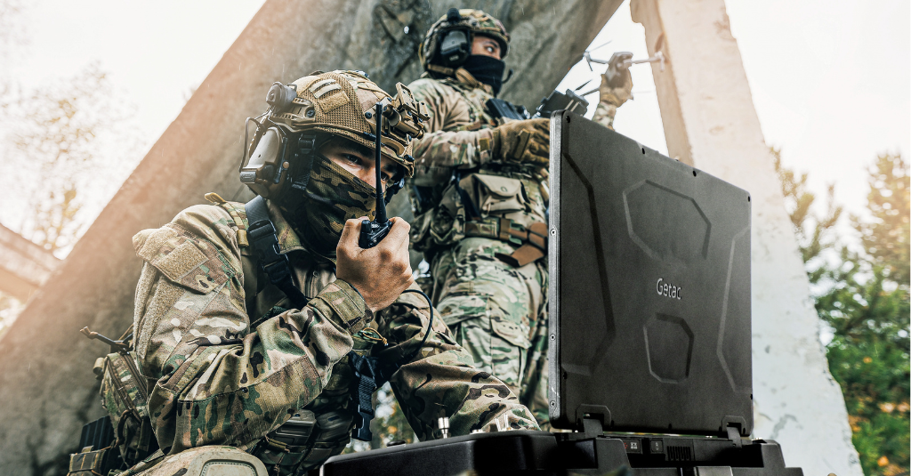 Soldiers in the field with a rugged Getac laptop, one using a handheld radio while another launches a small drone