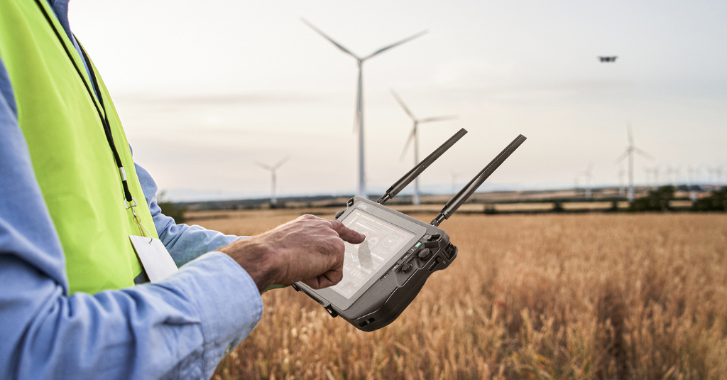 A utilities worker in a high-visibility vest using a Getac rugged handheld drone controller to inspect wind turbines in an open field, with a drone visible in the background.