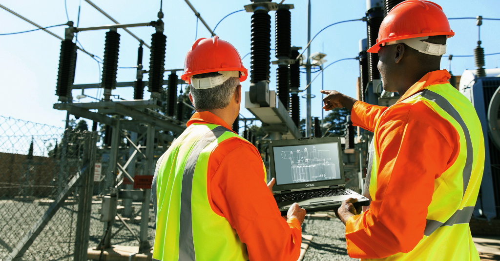 Two utility linemen in hard hats and hi-vis gear review electrical substation schematics on a rugged Getac laptop.