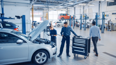 A person with a clipboard and mechanic tools standing in front of a car in a garage. The article provides tips for managing an automotive mechanic workshop effectively.