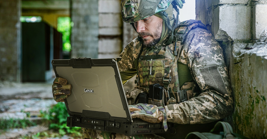 A soldier in camouflage uniform, helmet, and tactical gloves uses a Getac B360 Pro fully rugged laptop while crouched against a concrete wall in a field environment.