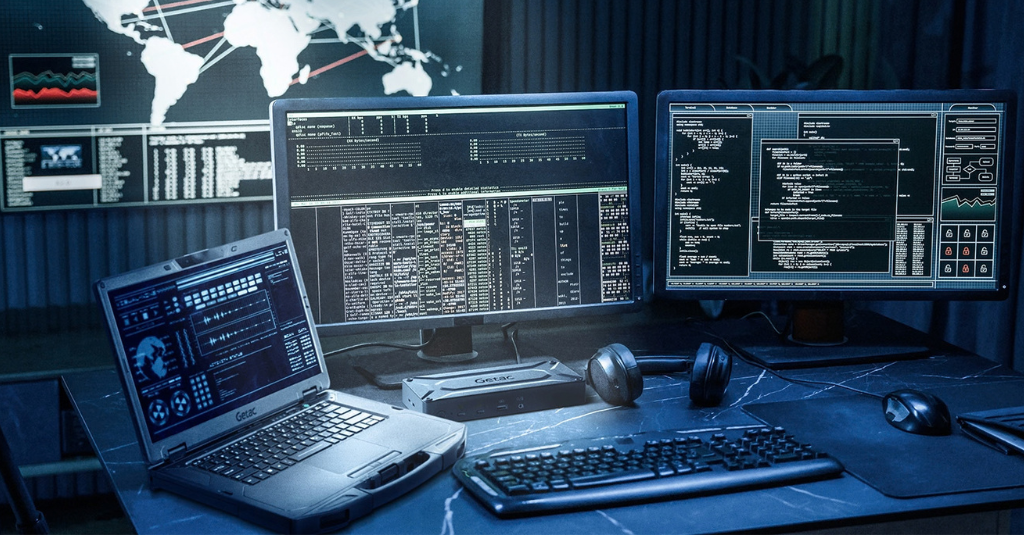 A Getac rugged laptop and a Getac docking device sit on a desk in a dimly lit operations center, next to two monitors showing data and code, with a world map display on the wall behind them.