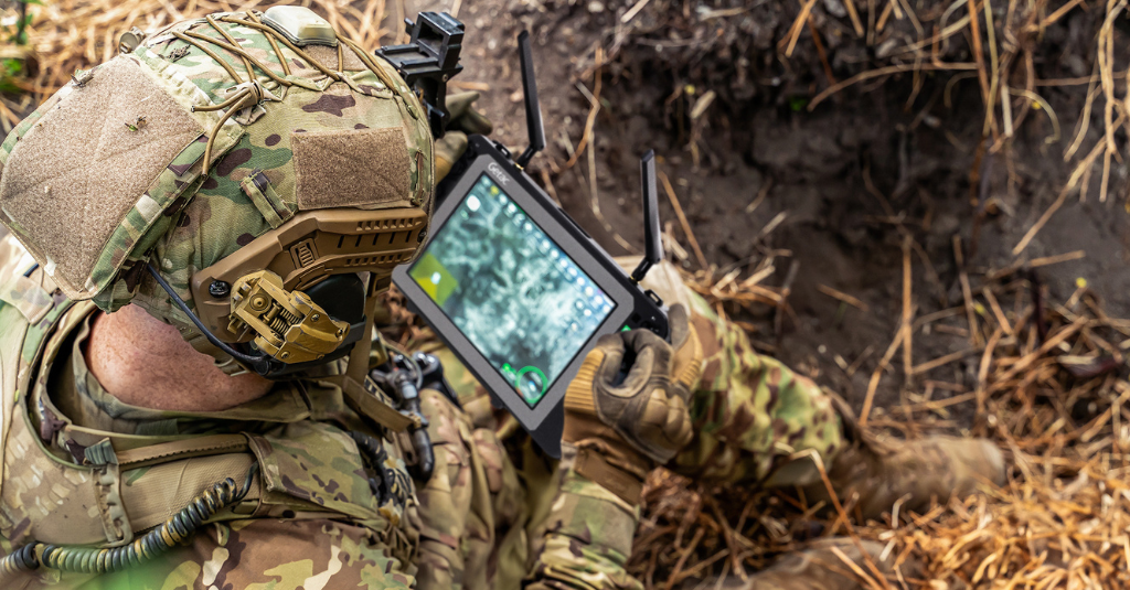 A soldier in full tactical gear using a Getac rugged tablet with an antenna attachment to control a drone in a field environment.