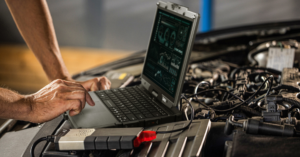 A technician uses a Getac rugged laptop displaying diagnostic dashboards while performing vehicle engine diagnostics under the hood, connected via cable to a diagnostic scan tool.