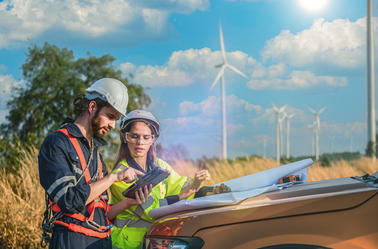 Two utility field engineers wearing safety gear review data on a rugged tablet beside a vehicle, with wind turbines in the background.
