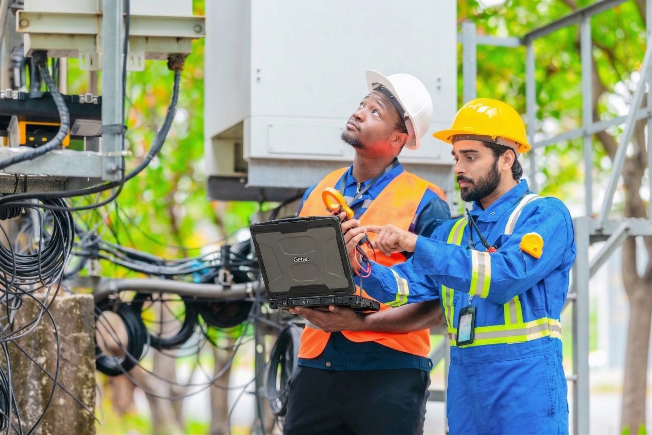 Telecom technicians using a Getac B360-fully-rugged laptop to inspect network infrastructure equipment in a field deployment environment