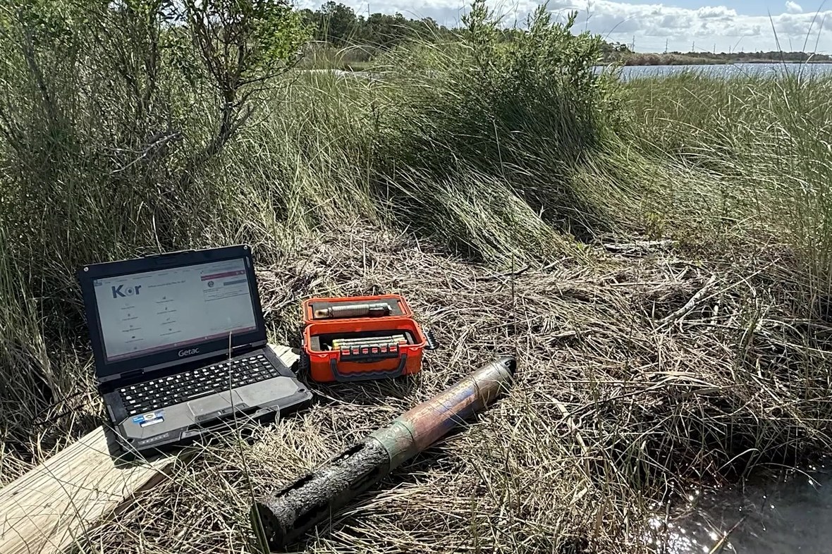 Getac B360 laptop beside a water quality sonde and its case in salt marsh grass at the water's edge